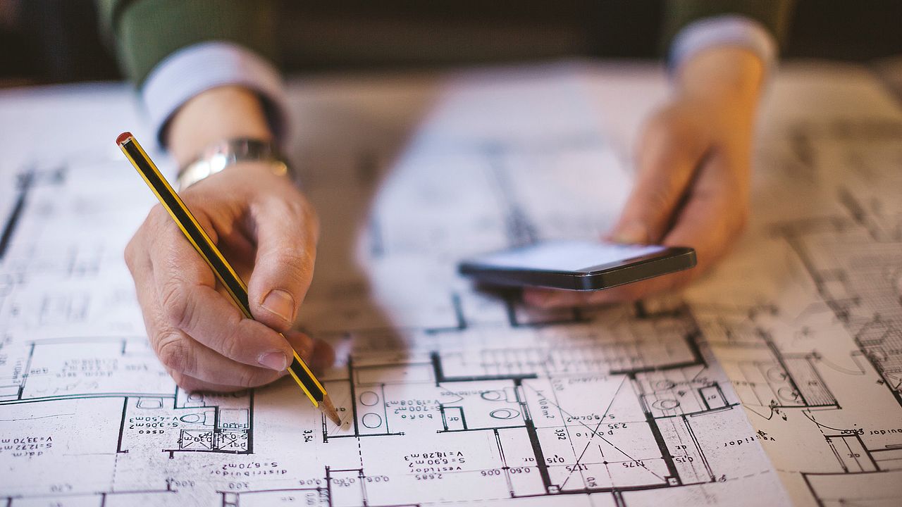 Detailaufnahme eines Bauplans, der von jemandem mit Stift und Smartphone studiert wird.