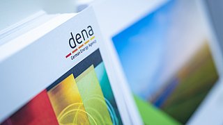 Das Bild zeigt mehrere Broschüren, darunter eine mit dem Logo 'dena' und dem Text 'German Energy Agency'. Die Broschüre hat eine bunte Gestaltung mit Farben in Rot, Gelb, Grün und Blau.
