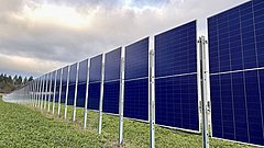 Reihe von blauen Solarpaneelen auf Metallgestellen in einer grünen Wiese unter bewölktem Himmel.