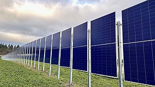 Reihe von blauen Solarpaneelen auf Metallgestellen in einer grünen Wiese unter bewölktem Himmel.