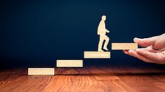 Eine Hand hält eine Treppe aus Holzblöcken, auf der eine stilisierte Figur nach oben steigt.