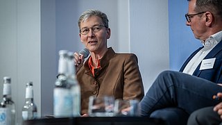 Zwei sitzende Personen bei einer Diskussion, eine trägt braune Jacke mit rotem Kragen, die andere einen dunkelblauen Anzug mit weißem Hemd.