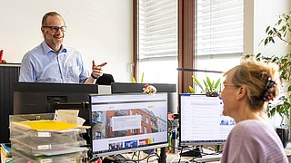 Ein Mann und eine Frau arbeiten in einem Büro, umgeben von Computern und Büromaterialien.