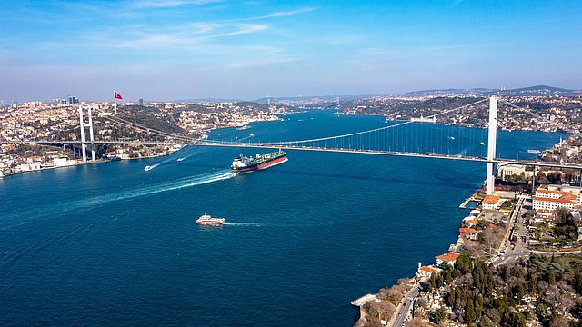 Luftaufnahme der Bosporus-Brücke mit einem Frachtschiff und einem kleinen Boot auf dem Wasser, umgeben von Hügeln und Stadtlandschaft.