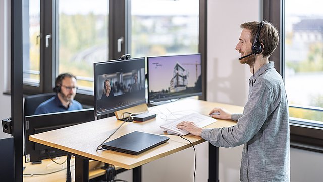 Zwei Personen mit Headset auf dem Kopf an ihren Arbeitsplätzen (Schreibtisch mit Bildschirmen, Notebook, Tastatur)