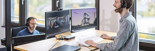 Zwei Personen mit Headset auf dem Kopf an ihren Arbeitsplätzen (Schreibtisch mit Bildschirmen, Notebook, Tastatur)