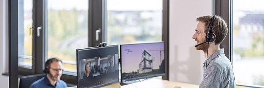 Zwei Personen mit Headset auf dem Kopf an ihren Arbeitsplätzen (Schreibtisch mit Bildschirmen, Notebook, Tastatur)