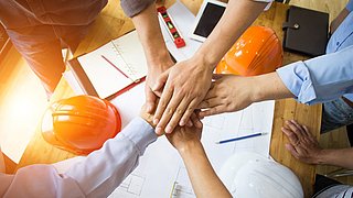 5 Hände übereinandergelegt, darunter 2 Bauhelme und Skizzen symbolisieren die Kraft der Zusammenarbeit im Baugewerbe.