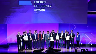 Gruppenszene von 14 Personen auf einer Bühne, die Urkunden für den Energy Efficiency Award 2022 halten. Im Hintergrund steht der Schriftzug 'ENERGY EFFICIENCY AWARD by dena'.