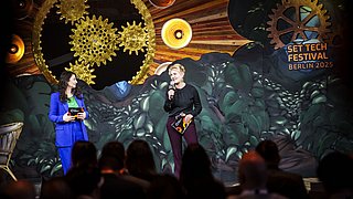 Zwei Moderatorinnen auf einer Bühne beim SET Tech Festival Berlin 2025, vor dekorativer Kulisse mit Zahnrädern und Pflanzen, Publikum im Vordergrund.