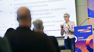 Frau mit kurzen grauen Haaren hält Präsentation vor Leinwand mit Text, steht neben Rednerpult mit Aufschrift 'Energiewende Kongress 2025 sicher vorangehen'.