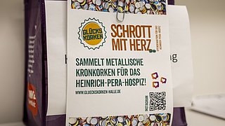 Ein lila Beutel mit einem Aufkleber. Der Aufkleber enthält den Text: 'GLÜCKSKORKEN SCHROTT MIT HERZ SAMMELT METALLISCHE KRONKORKEN FÜR DAS HEINRICH-PERA-HOSPIZ! WWW.GLUECKSKORKEN-HALE.DE'.