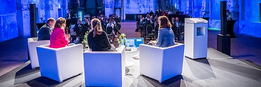 Von der Perspektive der Bühne aus sind vier Personen von hinten auf der Bühne auf Hockern sitzend zum Publikum gewidmet zu sehen.