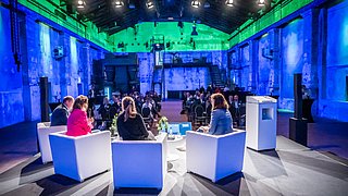 Von der Perspektive der Bühne aus sind vier Personen von hinten auf der Bühne auf Hockern sitzend zum Publikum gewidmet zu sehen.