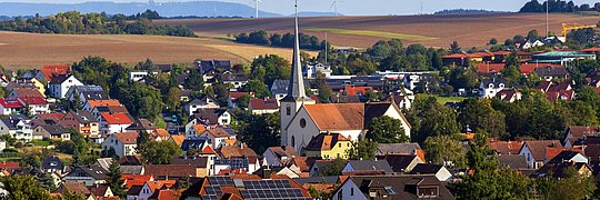 Dorf im Herbst mit einer Kirche und einem mit Solarmodulen ausgestatteten Haus im Zentrum
