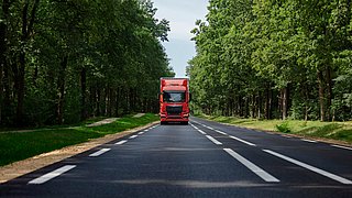 Red,Commercial,Truck,Traveling,On,Paved,Road,Through,Dense,Green