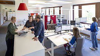 Mehrere Personen sind in einem Büro tätig, sie diskutieren und nutzen Computer für ihre Arbeit.