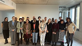 Gruppe von Menschen in einem modernen Raum mit Betonwand und großen Fenstern, verschiedene Kleidung und Posen.