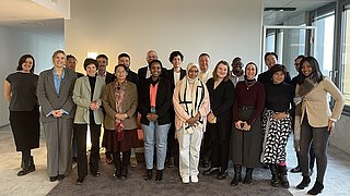 Gruppe von Menschen in einem modernen Raum mit Betonwand und großen Fenstern, verschiedene Kleidung und Posen.