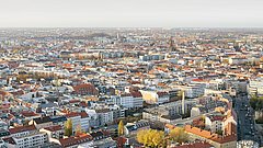 Panoramaansicht einer Stadt mit zahlreichen Gebäuden, roten Dächern und grünen Bäumen, unter einem klaren Himmel.