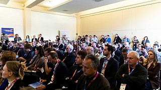 Blick ins Publikum, in dem viele Menschen sitzen und einige am Rand stehen. 