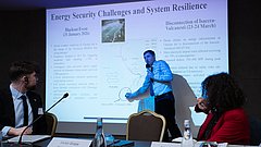 Mann in Hemd und Krawatte steht vor Projektionsleinwand und zeigt auf Diagramm, zwei Personen sitzen an Tisch mit Mikrofonen und Wasserflaschen und schauen ihn an.