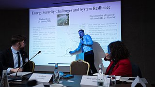 A man in a shirt and tie is standing in front of a projection screen, pointing at a diagram; two people are sitting at a table with microphones and water bottles, looking at him.