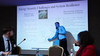 Mann in Hemd und Krawatte steht vor Projektionsleinwand und zeigt auf Diagramm, zwei Personen sitzen an Tisch mit Mikrofonen und Wasserflaschen und schauen ihn an.