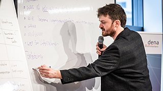 Ein Mann schreibt auf ein Whiteboard und spricht in ein Mikrofon.