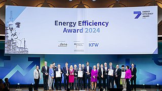 Gruppenfoto von Preisträgern des Energy Efficiency Award 2024 auf einer Bühne. Im Hintergrund steht ein großes Banner mit dem Text 'Energy Efficiency Award 2024' und Logos von dena und KFW. Die Personen halten Urkunden.