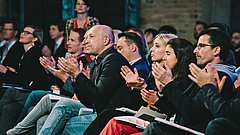 Eine Gruppe von Menschen sitzt auf Stühlen und applaudiert. Die meisten Personen sind in formeller Kleidung gekleidet. Einige halten Notizen in den Händen.