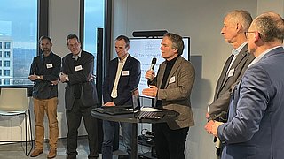 Fünf Männer in Businesskleidung stehen in einem Raum mit großen Fenstern, einer hält ein Mikrofon, ein Bildschirm zeigt den Schriftzug 'Akteurstreffen'.