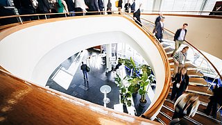 Menschen bewegen sich auf einer geschwungenen Treppe mit Holzgeländer in einem modernen Gebäude mit großen Fenstern und Pflanzen im Innenraum.