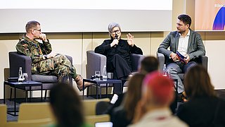 Drei Personen sitzen auf Sesseln auf einer Bühne, eine trägt eine Militäruniform, die anderen zwei sind in Zivilkleidung, Publikum im Vordergrund unscharf.