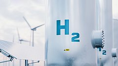 Nahaufnahme eines silbernen Wasserstofftanks mit der Aufschrift 'H2'. Im Hintergrund sind Windturbinen und Solarpanels sichtbar.