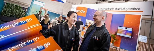 Zwei Personen in schwarzen Jacken mit dem Logo "dena" stehen vor einem Messestand mit Bannern und Bildschirmen, auf denen "Energie to go" und "dena.de" zu sehen sind.