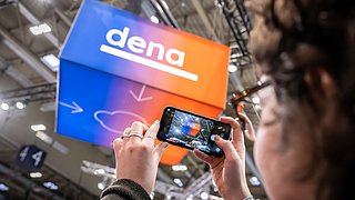 Person hält Smartphone hoch und fotografiert einen blau-orangefarbenen Würfel mit dem Schriftzug 'dena' in einer Halle