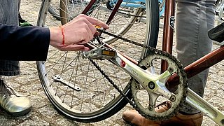 Eine Hand hält einen Schraubenschlüssel und arbeitet an der Kette eines Fahrrads. Im Hintergrund sind weitere Fahrräder und Werkzeuge sichtbar.