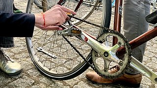 Eine Hand hält einen Schraubenschlüssel und arbeitet an der Kette eines Fahrrads. Im Hintergrund sind weitere Fahrräder und Werkzeuge sichtbar.