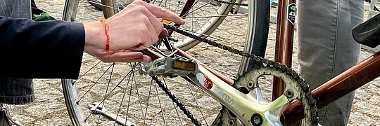 Eine Hand hält einen Schraubenschlüssel und arbeitet an der Kette eines Fahrrads. Im Hintergrund sind weitere Fahrräder und Werkzeuge sichtbar.