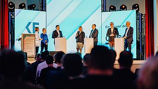 Sechs Personen stehen auf einer Bühne vor großen Bildschirmen und sprechen vor einem Publikum