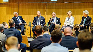 Fünf Personen in Anzügen und formeller Kleidung sitzen auf einer Bühne vor einem Publikum, eine Person spricht in ein Mikrofon