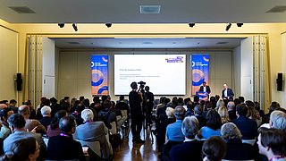 Blick auf das Podium im Hintergrund, das Publikum ist im Vordergrund zu sehen. 