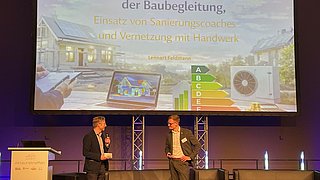 Zwei Männer in Anzügen stehen vor einer Bühne mit Sofas und einem großen Bildschirm, der eine Präsentation zu rechtlichen Grundlagen der Baubegleitung zeigt