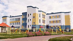 Mehrstöckiges Gebäude mit gelben und blauen Akzenten, umgeben von einem Spielplatz mit Spielgeräten und Bänken im Vordergrund.