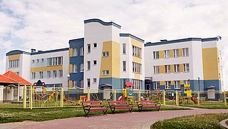 Mehrstöckiges Gebäude mit gelben und blauen Akzenten, umgeben von einem Spielplatz mit Spielgeräten und Bänken im Vordergrund.