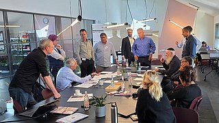 Gruppe von Menschen in einem modernen Besprechungsraum um einen langen Tisch versammelt, einige sitzen, andere stehen, mit Laptops, Getränken und Unterlagen auf dem Tisch