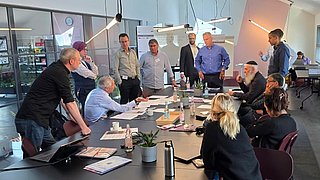 Gruppe von Menschen in einem modernen Besprechungsraum um einen langen Tisch versammelt, einige sitzen, andere stehen, mit Laptops, Getränken und Unterlagen auf dem Tisch