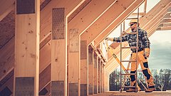 Ein Handwerker steht auf einer Leiter und misst Holzrahmen in einem Dachstuhl. Er trägt eine gelbe Mütze, eine kariierte Jacke und orangefarbene Arbeitshosen.