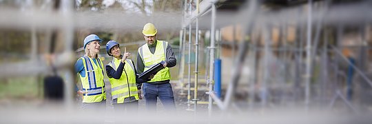 Drei Personen mit Schutzhelmen und Warnwesten stehen auf einer Baustelle, eine Person zeigt mit dem Finger nach oben, eine hält eine Bauzeichnung